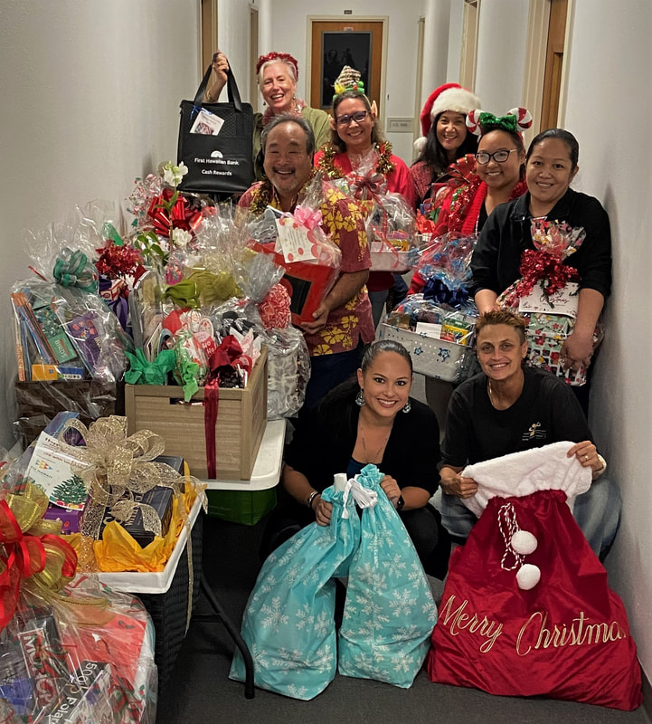 Members of the Hawaii Chamber of Commerce in festive holiday gear together with Going Home Hawaii staff in black GHH t-shirts hold gift baskets and bags beautifully decorated with shiny bows, holiday gift paper or bags, and full of enticing items. 